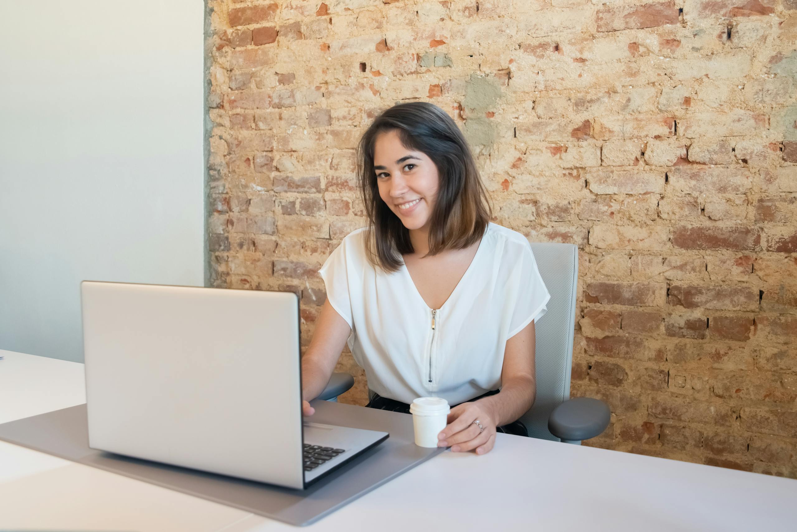 A cheerful woman working on a laptop in a rustic office setting, holding a cup.