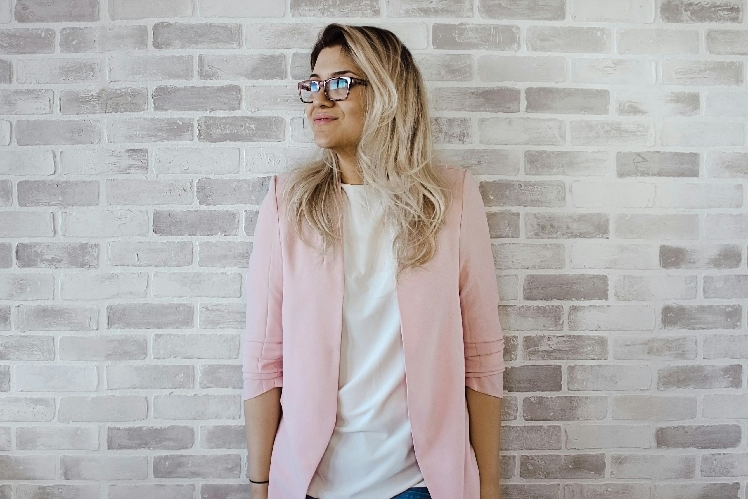 Elegant woman with glasses poses against a brick wall in a trendy pink blazer.