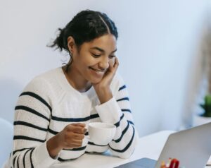 Positive young African American female freelancer in casual clothes talking on smartphone and drinking coffee while working remotely on laptop at home