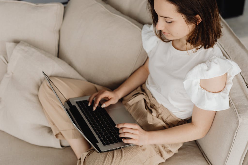 Young woman engaged in remote work on a laptop, sitting on a beige couch indoors.