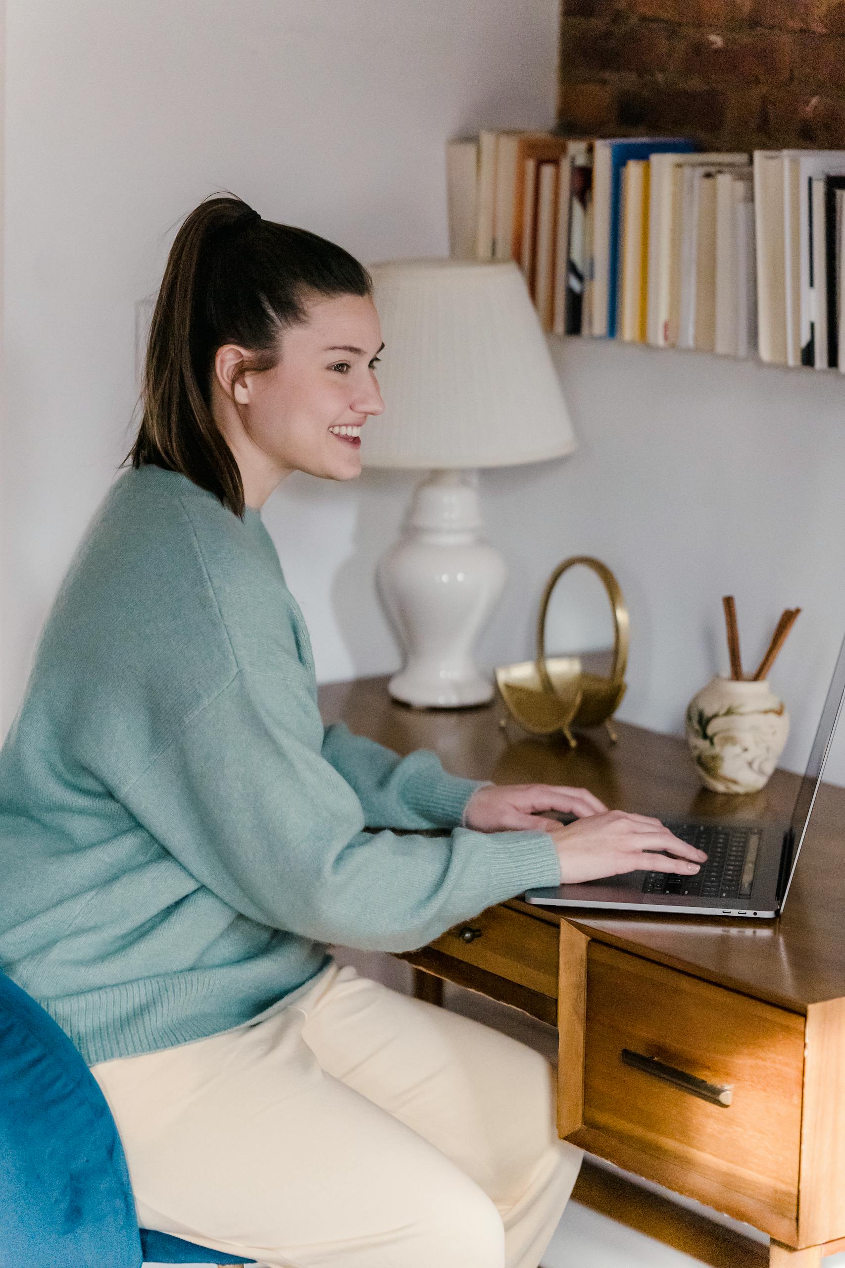 Side view of smiling female sitting at wooden desk with laptop and looking away while working online