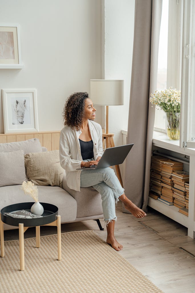 African American woman working on laptop at home, enjoying natural light by the window.