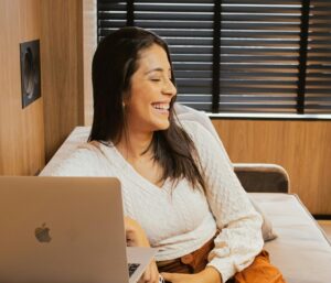 Caucasian woman on sofa in Brazil, using laptop and camera, smiling happily.