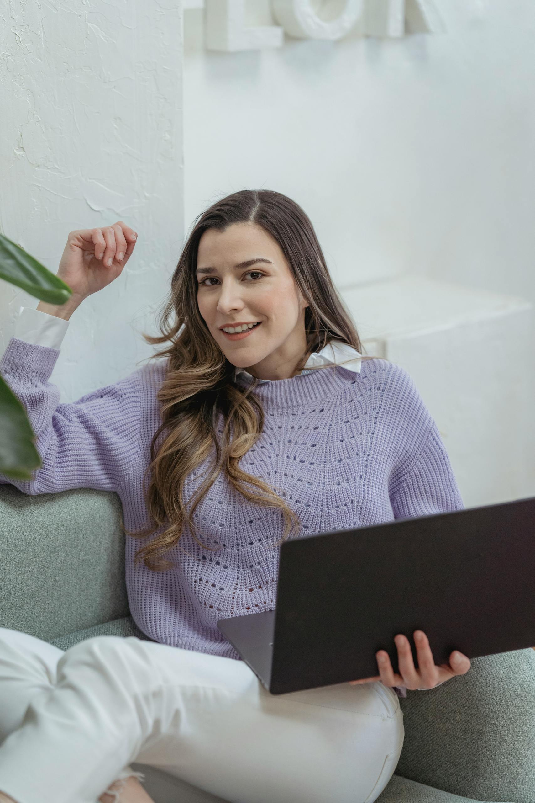 Positive young female freelancer with long wavy hair in stylish sweater smiling and looking at camera while sitting in armchair and working remotely on laptop