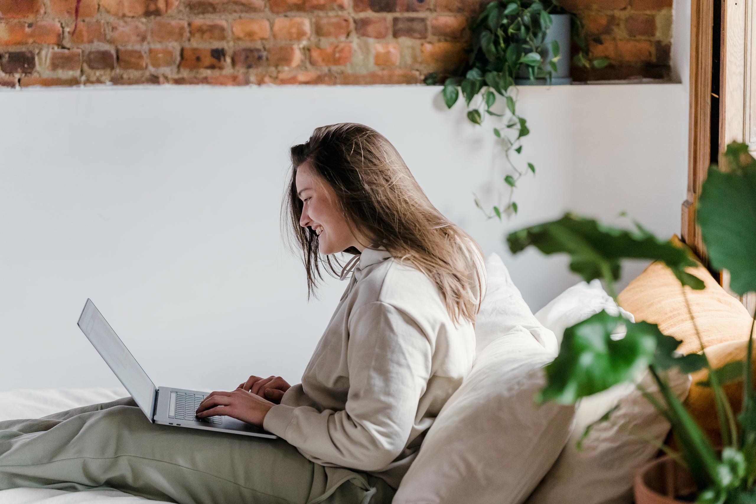 Side view of positive female typing on modern netbook while sitting on comfortable bed near brick wall in room with green potted plants