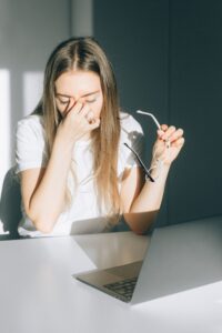 Young woman holds her glasses, experiencing digital eye strain in a bright office.