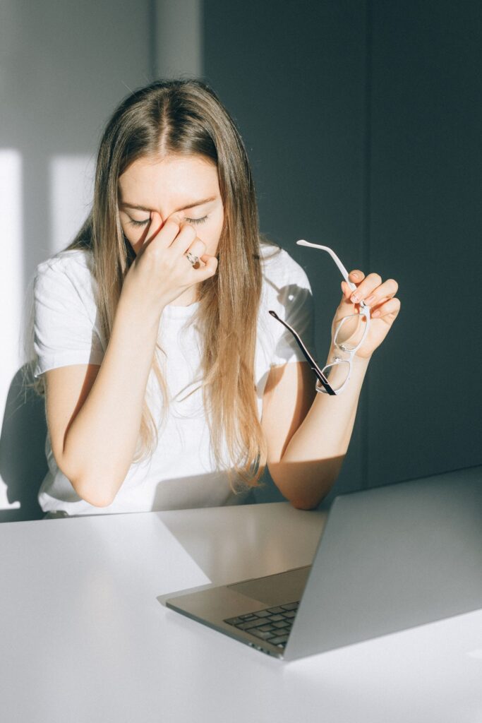Young woman holds her glasses, experiencing digital eye strain in a bright office.